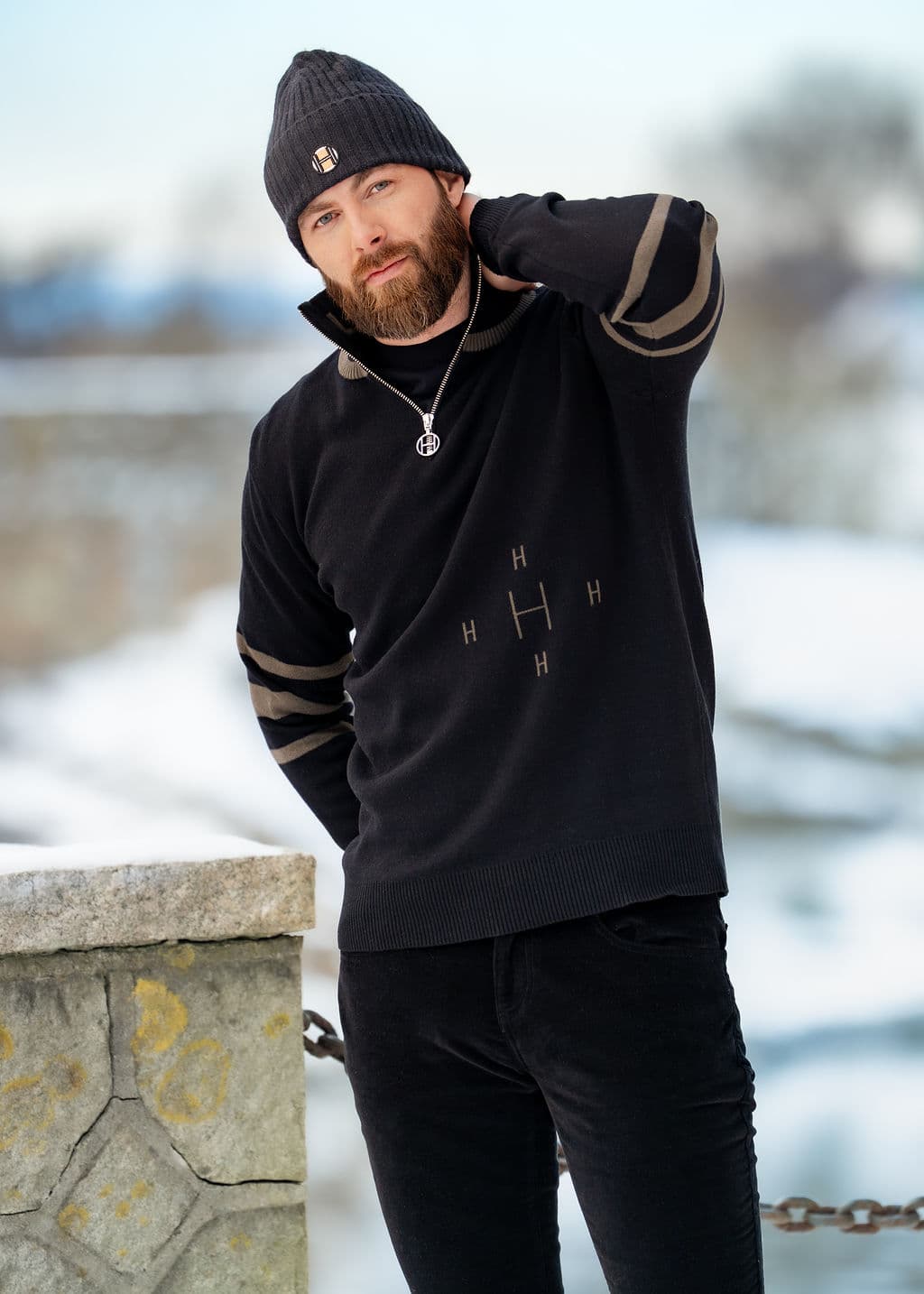 Man wearing a dark sweater and beanie, standing outdoors in a snowy landscape, with one hand on his neck and leaning against a stone wall.