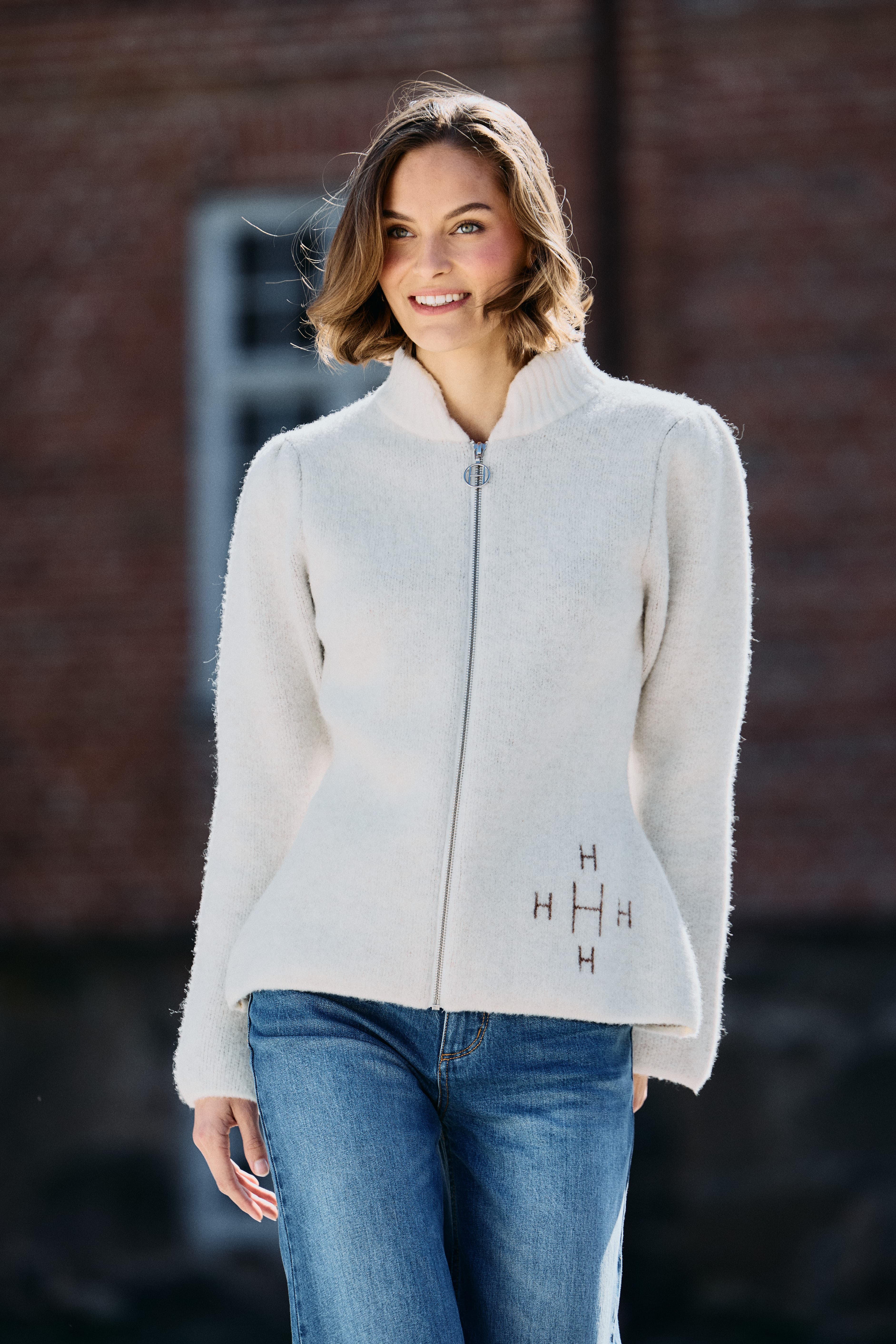 A smiling woman wearing a cream zip-up jacket with embroidery and blue jeans, standing outdoors against a brick building background.