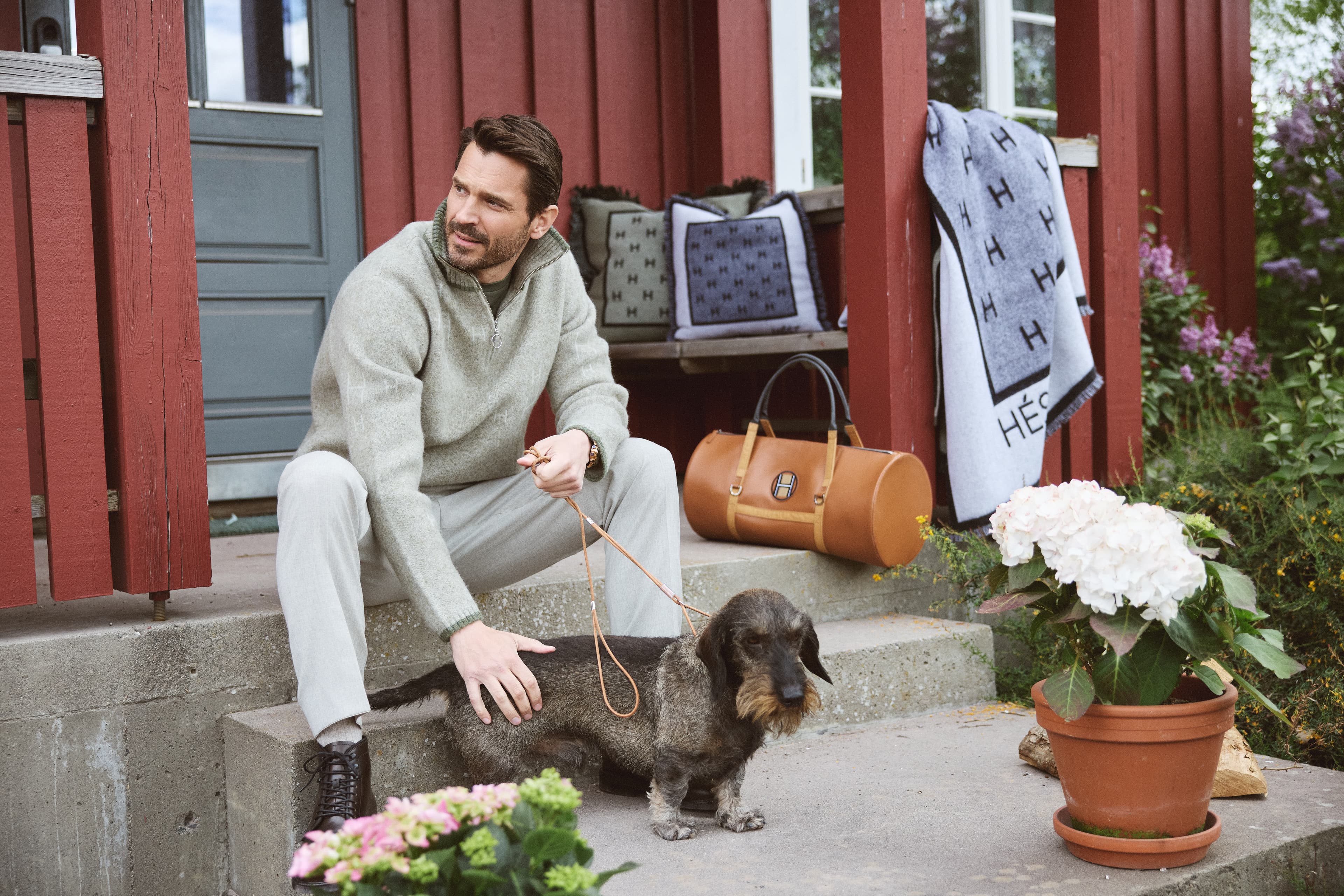 Man sitting on steps petting a dog, surrounded by potted flowers and bags, in front of a red building.