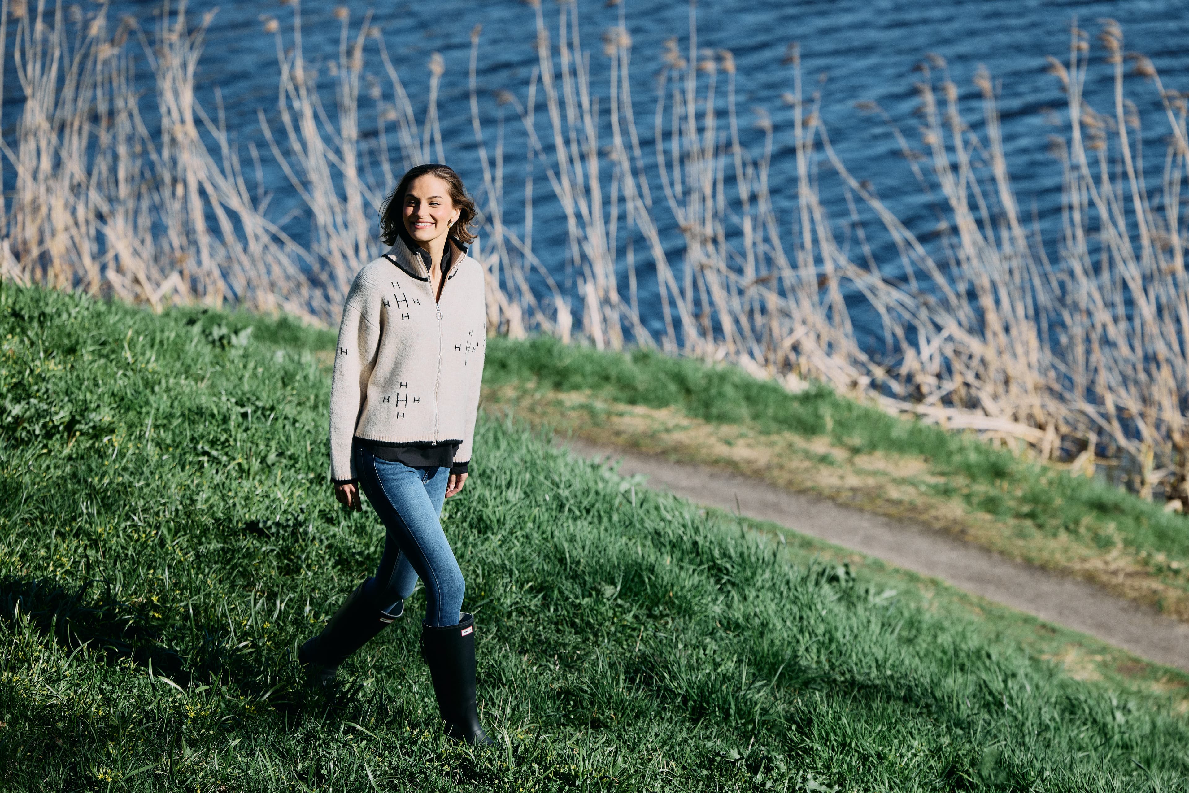 A woman in a light sweater and jeans walks on a grassy path by a lakeside, with tall reeds and water in the background on a sunny day.