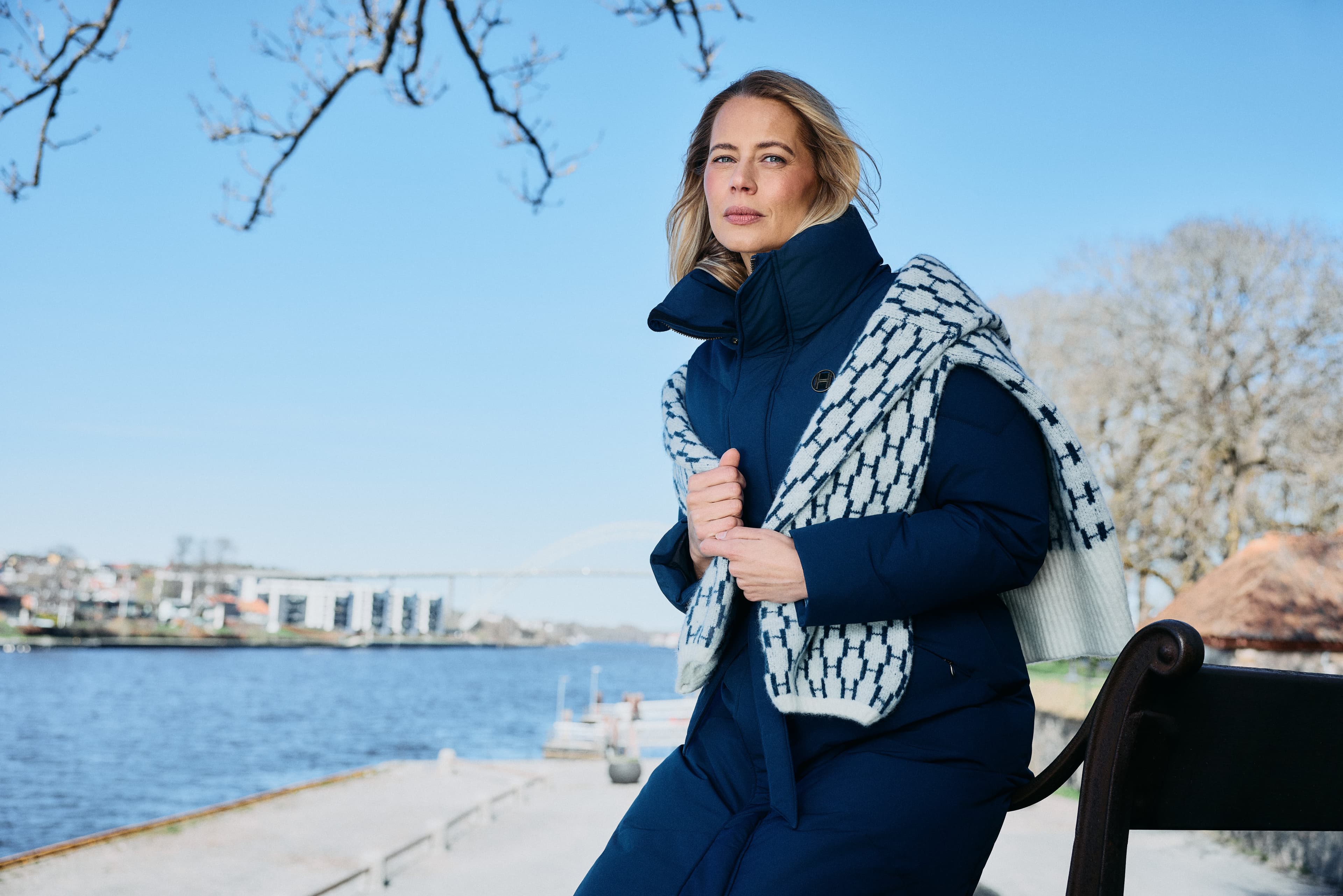 A person in a blue coat and knittet sweater over the shoulders stands near a waterfront, with a bridge and buildings in the background on a clear day.