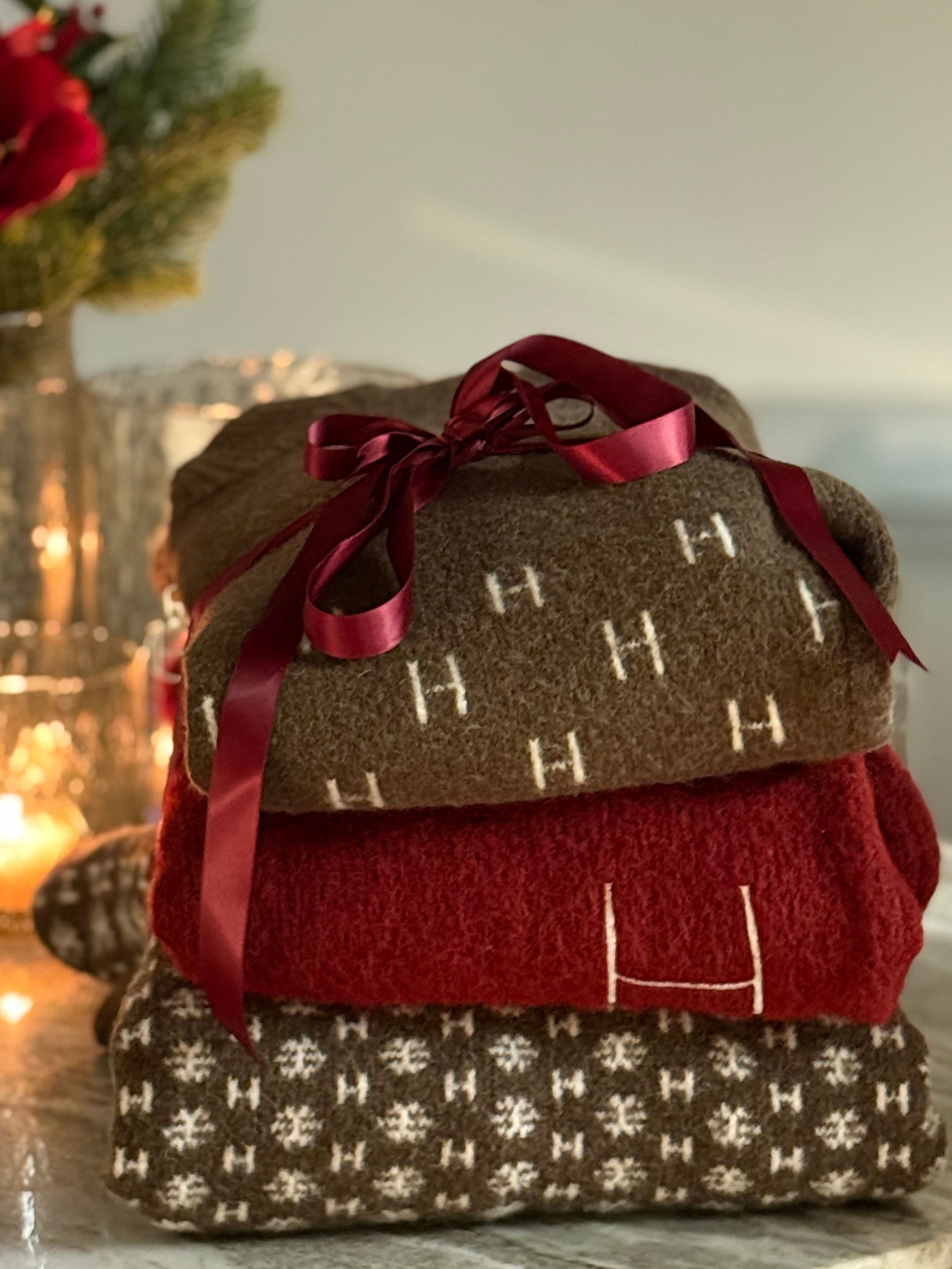 Three folded knit sweaters (brown, red, patterned) tied with a burgundy ribbon, candlelight and evergreen decor blurred in the background.